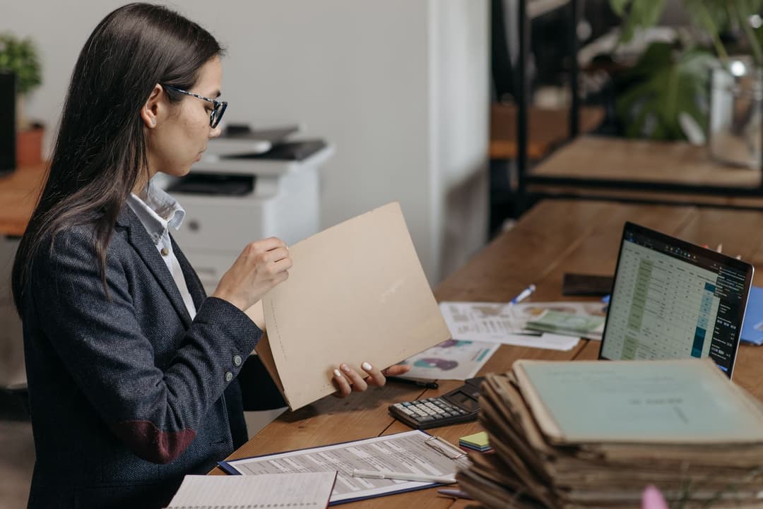 Eine Büromitarbeiterin sortiert Mappen, neben ihr ein Stapel Ordner und ein Laptop